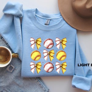 May include: Light blue sweatshirt featuring a softball and bow design. The softball is yellow with red stitching and the bows are white with yellow accents. The text "LIGHT BLUE" is printed on the bottom right corner of the sweatshirt.