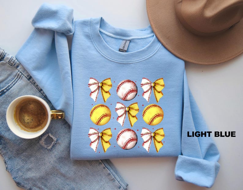 May include: Light blue sweatshirt featuring a softball and bow design. The softball is yellow with red stitching and the bows are white with yellow accents. The text "LIGHT BLUE" is printed on the bottom right corner of the sweatshirt.