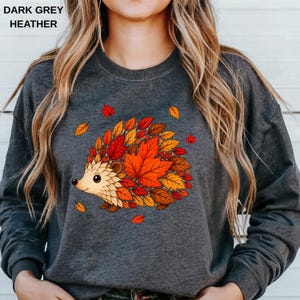 May include: A dark grey heather crewneck sweatshirt featuring a graphic of a hedgehog whose back is composed of vibrant red, orange, and yellow autumn maple leaves. The text "DARK GREY HEATHER" is printed in the upper left corner of the image.
