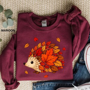May include: A maroon long-sleeve sweatshirt featuring a graphic illustration of a hedgehog whose back is covered in vibrant autumn leaves in shades of orange, red, and brown. The sweatshirt has a crew neckline and a visible brand label at the collar.