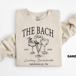 May include: A beige sweatshirt with the text "THE BACH Club EST. 2025" and a graphic of three cocktail glasses. The text "Lindsey Bachelorette NASHVILLE, TN" is below the graphic. The word "SAND" is in the bottom right corner of the image.