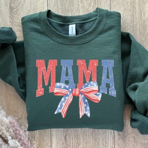 Pu&ograve; includere: Una felpa verde scuro con la parola "MAMA" stampata in rosso, bianco e blu con un fiocco rosso, bianco e blu.