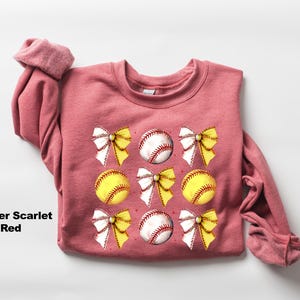 May include: Heather scarlet red sweatshirt with a graphic design of white bows and yellow and white baseballs.