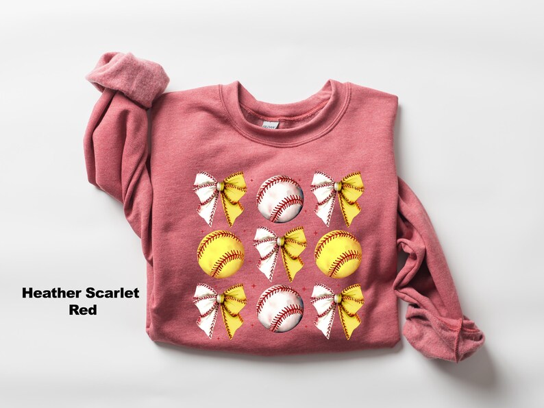 May include: Heather scarlet red sweatshirt with a graphic design of white bows and yellow and white baseballs.