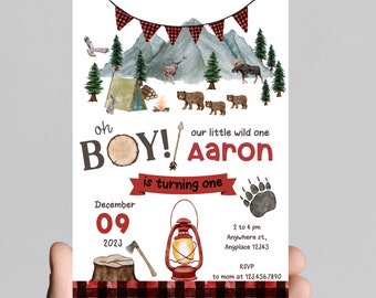 May include: A woodland-themed first birthday party invitation with a red and black plaid background. The invitation features a watercolor illustration of a mountain scene with a tent, campfire, and animals. The text reads "Oh Boy! Our little wild one Aaron is turning one. December 09, 2023. 2 to 4 pm. Anywhere st, Anyplace 12345. RSVP to mom at 123.456.7890."