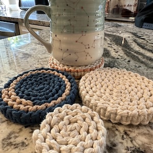May include: Four crocheted coasters in beige and blue. The coasters are round and have a textured pattern. A mug with a constellation design is sitting on top of one of the coasters.