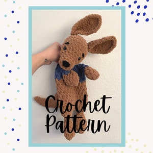 Puede incluir: Patrón de crochet para un perro de peluche marrón con ropa azul y marrón. El perro está siendo sostenido por una mano con un pequeño tatuaje.
