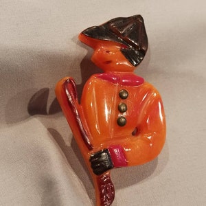 May include: Orange and black plastic brooch of a person wearing a hat and holding a stick. The person is wearing a red and orange jacket with gold buttons.