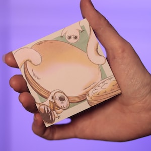 Puede incluir: Una tarjeta cuadrada con una ilustración caprichosa de una criatura con ojos grandes y un cuerpo redondo. La obra de arte tiene una paleta de colores suaves con acentos beige, marrón y verde claro. La tarjeta se sostiene sobre un fondo púrpura.