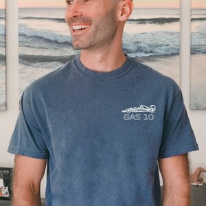May include: A man wearing a blue t-shirt with a white graphic of a race car and the text "GAS 10". He is standing in front of a wall with three framed pictures of a beach scene.