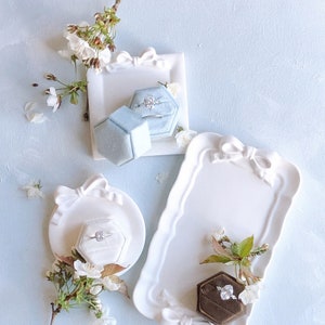 May include: Three white ceramic ring dishes with a bow design. Each dish has a small velvet ring box inside. The boxes are blue, brown, and white. The dishes are on a light blue background with white flowers.