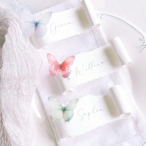 May include: Three white paper place cards with torn edges, each featuring a watercolour butterfly and a name written in cursive script. The top card reads Groom, the middle card reads William, and the bottom card reads Sophia. The cards are rolled at the ends.