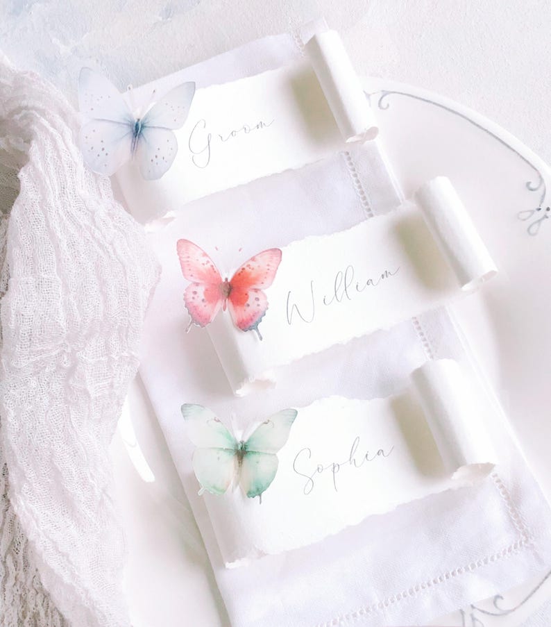 May include: Three white paper place cards with torn edges, each featuring a watercolour butterfly and a name written in cursive script. The top card reads Groom, the middle card reads William, and the bottom card reads Sophia. The cards are rolled at the ends.