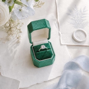 May include: A silver ring with a central round-cut stone and smaller side stones, displayed inside an open octagonal forest green velvet ring box with a white interior lining, positioned on a textured white paper surface.