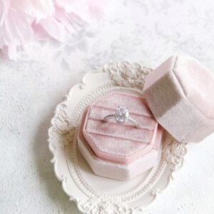 May include: A silver ring with an oval-cut center stone and a pav&eacute; band, resting inside an octagonal light pink velvet ring box. The box is placed on a small, oval, cream-colored decorative tray with an ornate, raised floral border design.