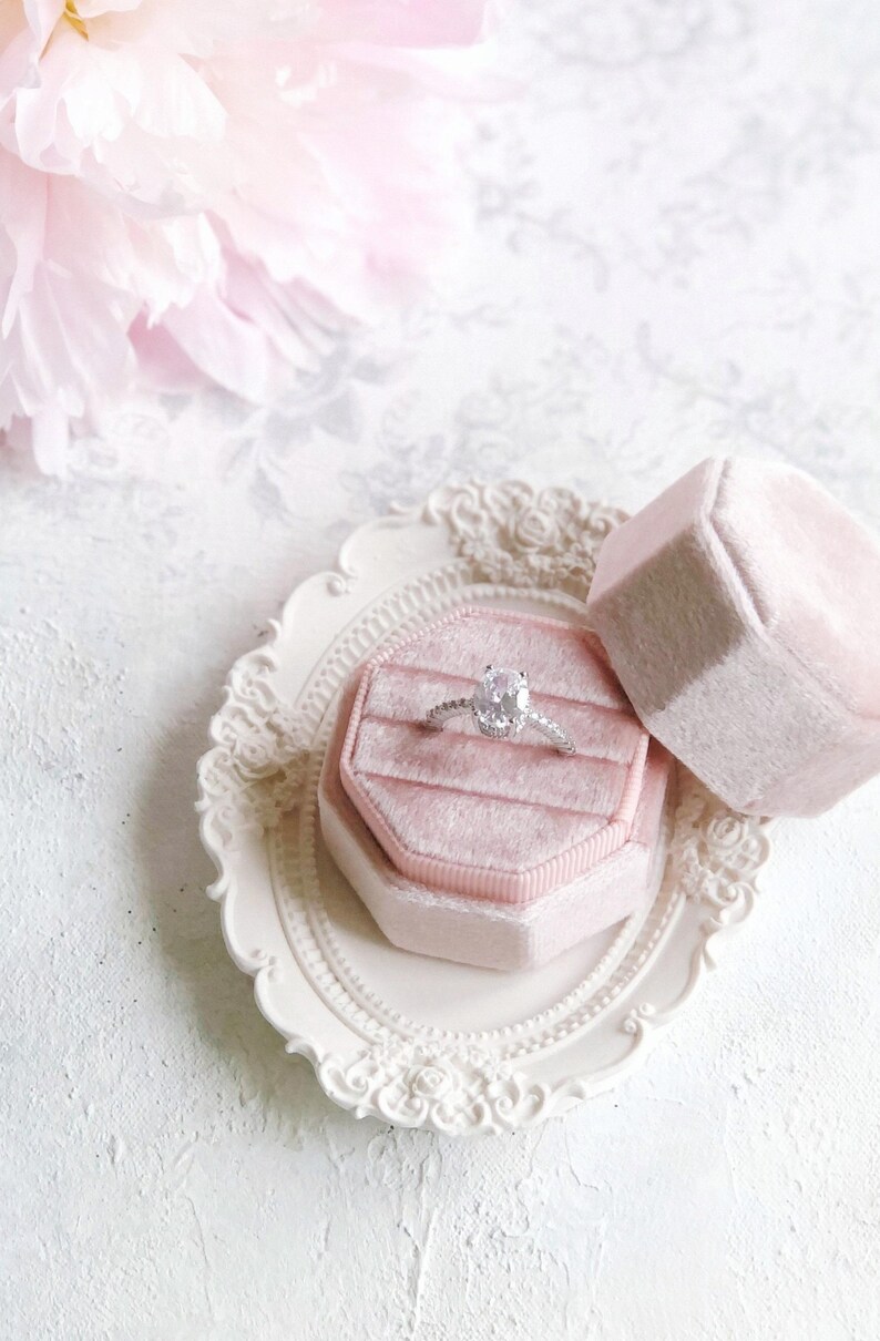 May include: A silver ring with an oval-cut center stone and a pav&eacute; band, resting inside an octagonal light pink velvet ring box. The box is placed on a small, oval, cream-colored decorative tray with an ornate, raised floral border design.