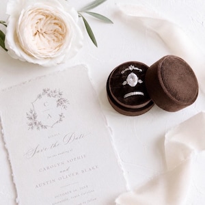 May include: A brown velvet ring box containing three silver-toned rings, positioned next to a cream-colored Save the Date card with deckled edges. The card features the text Carolyn Sophie and Austin Oliver Blake, October 26, 20XX.