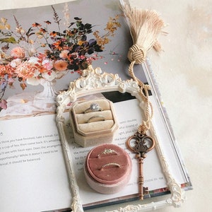 K&ouml;nnte beinhalten: Ein wei&szlig;er, verzierter Rahmen mit einer Samtringbox im Inneren. Die Ringbox ist rosa und enth&auml;lt einen goldenen Ring. Der Rahmen liegt auf einem Buch mit einem Blumendesign. Der Text auf dem Buch lautet "semi-double, eine volle Lichterkette, plus eine H&auml;lfte, die Basis von wie jetzt"