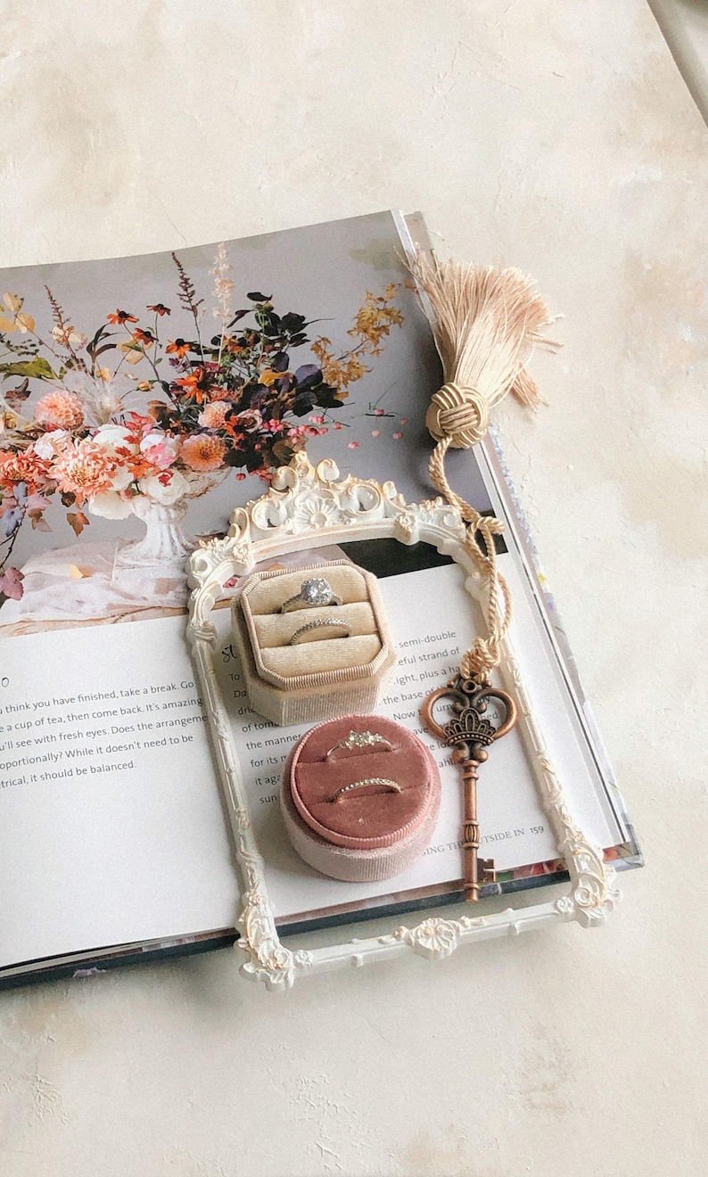 K&ouml;nnte beinhalten: Ein wei&szlig;er, verzierter Rahmen mit einer Samtringbox im Inneren. Die Ringbox ist rosa und enth&auml;lt einen goldenen Ring. Der Rahmen liegt auf einem Buch mit einem Blumendesign. Der Text auf dem Buch lautet "semi-double, eine volle Lichterkette, plus eine H&auml;lfte, die Basis von wie jetzt"