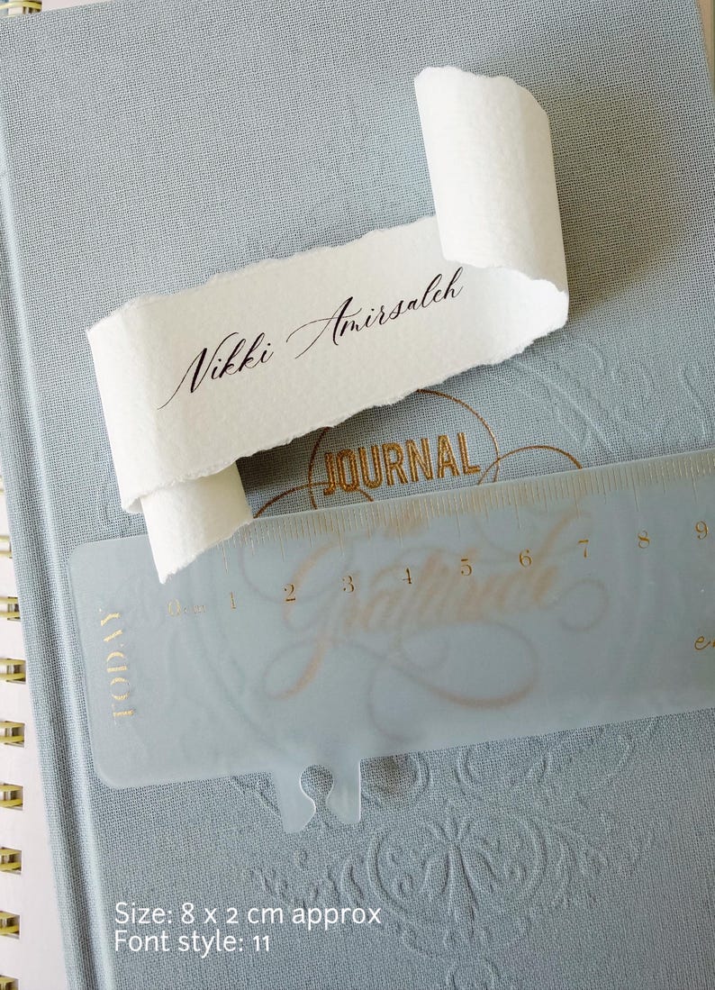 May include: A light blue journal with gold embossed text and a translucent plastic ruler showing a measurement of 8 cm by 2 cm. A white paper scroll with deckled edges features the black printed name Nikki Amirsaleh in script font.