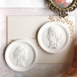May include: Two white, circular wax seals with classical female profile reliefs. One seal features a woman with a beaded necklace and a crown. The other seal shows a woman with a laurel wreath. The seals are on a cream-coloured card.