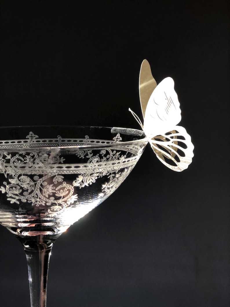 Puede incluir: Una copa de c&oacute;ctel de cristal transparente con un intrincado patr&oacute;n floral blanco grabado en el borde. Una mariposa de papel blanco con el nombre Emily impreso en cursiva est&aacute; sujeta al borde de la copa sobre un fondo negro.