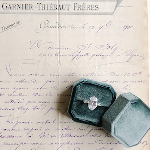 Op de afbeelding: Een vintage brief met blauwe inkt geschreven op crèmekleurig papier. De brief is dubbelgevouwen en heeft een blauwe fluwelen ringdoos met een diamanten ring erop. De brief luidt "Gerardmer, Vosges, le 17.9. 1902. Monsieur J. Joly, 57,57 Cours Lafayette prolongé à Lyon."
