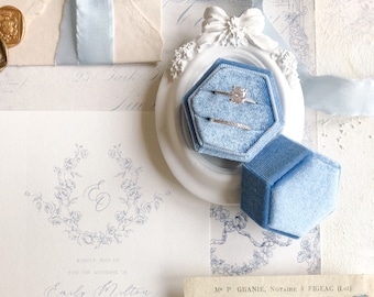 May include: A hexagonal light blue velvet ring box containing two silver-toned rings with clear stones, resting on a white oval frame with a bow detail, positioned next to a wedding invitation with floral illustrations and the names Emily Milton and Oliver Frank.