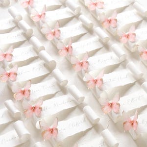 Könnte beinhalten: Eine Ansammlung von weißen Papierrollen mit gerissenen Kanten, die jeweils handgeschriebene Namen in grauer Tinte tragen und mit einem kleinen, zarten, durchscheinenden rosa Schmetterling verziert sind. Die Rollen sind in einem Raster angeordnet.