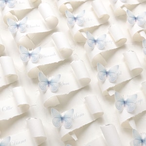 May include: A collection of white paper scrolls with torn edges, each featuring a light blue butterfly accent and a printed name. The names include Claudia, Hannah, Sara, Ollie, Diana, Alex, and Maria, displayed in a repeating pattern.