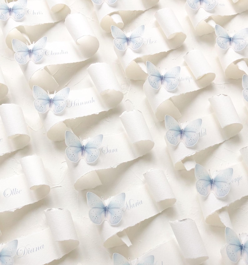 May include: A collection of white paper scrolls with torn edges, each featuring a light blue butterfly accent and a printed name. The names include Claudia, Hannah, Sara, Ollie, Diana, Alex, and Maria, displayed in a repeating pattern.