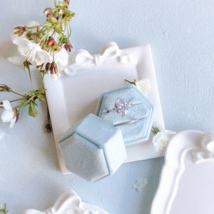 Puede incluir: Una caja de anillo de terciopelo azul claro con forma hexagonal. La caja está abierta y un anillo de plata con un gran diamante de forma ovalada está dentro. La caja de anillo está sentada sobre una superficie blanca con un lazo blanco.