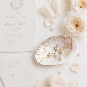 May include: A cream-colored wedding invitation suite with deckled edges and gold lettering, featuring a pair of gold-toned drop earrings with white pearls and clear crystals resting inside an iridescent seashell, surrounded by white roses and pearls.