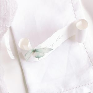 May include: A white paper scroll with deckled edges featuring the name Sophia in cursive script. A delicate, pale green butterfly embellishment is attached to the left side of the scroll, which rests on a white fabric napkin with a hemstitched border.