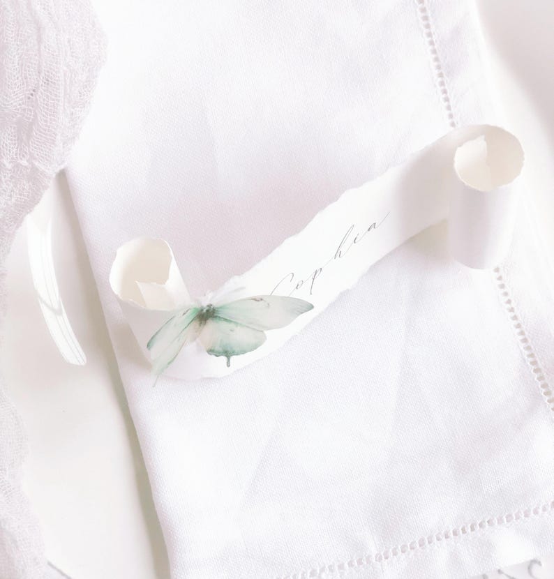 May include: A white paper scroll with deckled edges featuring the name Sophia in cursive script. A delicate, pale green butterfly embellishment is attached to the left side of the scroll, which rests on a white fabric napkin with a hemstitched border.