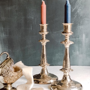 Op de afbeelding: Twee sierlijke zilveren kandelaars met brandende kaarsen, één koraal en één blauw. Een zilveren decoratieve container met bloemendetails en een crèmekleurig lint zijn ook aanwezig. De kandelaars hebben een gedetailleerde basis en steel.