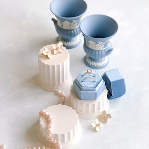 May include: Three white ceramic ring and earring display stands with blue and white ceramic urns, a blue velvet ring box, and pearl necklace. The stands have a fluted design and are decorated with delicate floral accents.