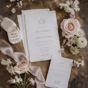 Könnte beinhalten: Elegante Hochzeitseinladungssuite mit einer Hauptkarte, einer RSVP-Karte und einer Samt-Ringbox mit Ringen. Die Schreibwaren haben einen Büttenrand und sind mit floralen Akzenten verziert, darunter Rosen und zarte Blüten. Ein Seidenband sorgt für einen Hauch von Luxus.