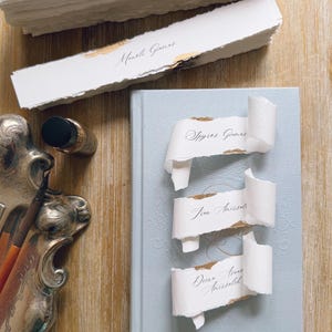 May include: A stack of white deckle-edge paper place cards with gold leaf accents and black calligraphy names, including Andrea Ortiz Carregha and Manoli Gianas, resting near a light blue hardcover book with three curled place cards.