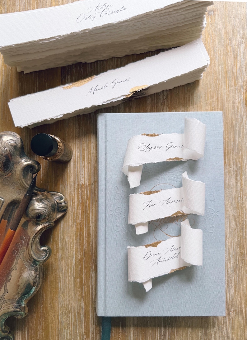 May include: A stack of white deckle-edge paper place cards with gold leaf accents and black calligraphy names, including Andrea Ortiz Carregha and Manoli Gianas, resting near a light blue hardcover book with three curled place cards.