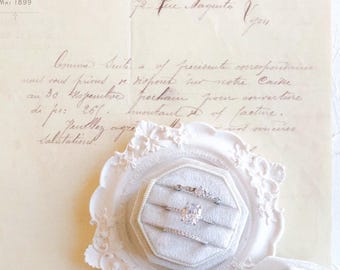 May include: Three silver-toned rings with clear stones are displayed in a cream-colored velvet ring box with an ornate, scalloped edge. The box rests on a piece of vintage paper featuring handwritten French cursive text and printed headers.