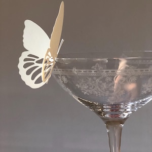 Puede incluir: Una copa de cristal transparente con un patr&oacute;n floral grabado alrededor del borde, que presenta una mariposa de papel blanco troquelada sujeta al borde. La mariposa tiene intrincados recortes en las alas y est&aacute; colocada verticalmente.