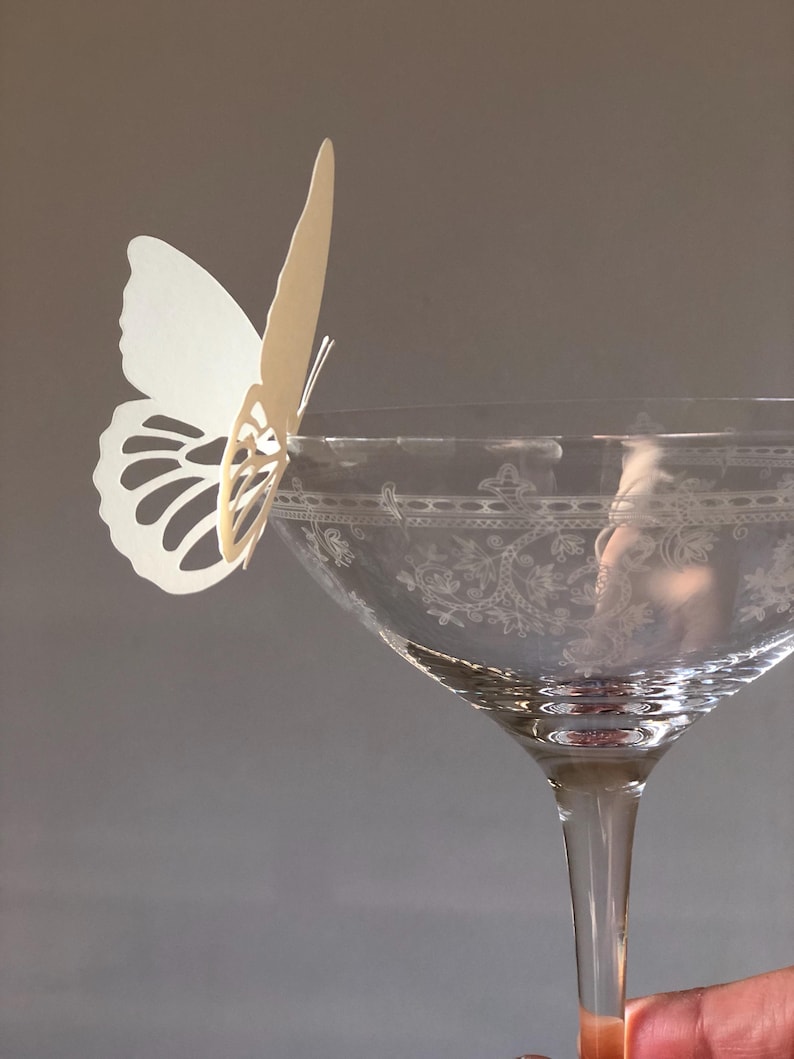 Puede incluir: Una copa de cristal transparente con un patr&oacute;n floral grabado alrededor del borde, que presenta una mariposa de papel blanco troquelada sujeta al borde. La mariposa tiene intrincados recortes en las alas y est&aacute; colocada verticalmente.