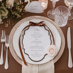 May include: A white scalloped menu card with dark brown edges, tied with a brown satin ribbon and adorned with a pale pink rose. The menu is placed on a white plate, a woven charger, and a brown tablecloth, accompanied by silver cutlery.