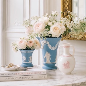 May include: Two blue ceramic vases with white relief designs and a white ceramic vase with pink relief designs. The vases hold bouquets of white and pink roses and other flowers. Books and a small plate are also on the marble surface.