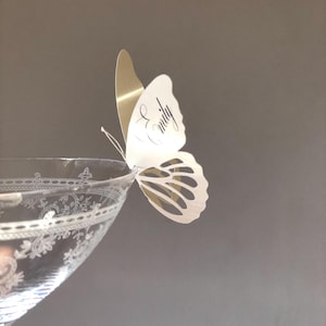 Puede incluir: Un vaso transparente con un patr&oacute;n floral grabado presenta una mariposa de papel blanca y dorada posada en el borde. La mariposa tiene el nombre Emily escrito en cursiva negra en el ala superior y detalles recortados en el ala inferior.