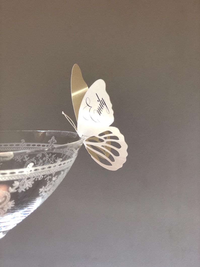 Puede incluir: Un vaso transparente con un patr&oacute;n floral grabado presenta una mariposa de papel blanca y dorada posada en el borde. La mariposa tiene el nombre Emily escrito en cursiva negra en el ala superior y detalles recortados en el ala inferior.