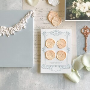 K&ouml;nnte beinhalten: Vier pfirsichfarbene Wachssiegel mit einem floralen Design sind auf einer wei&szlig;en Karte mit einem blauen und wei&szlig;en Rand angeordnet. Die Karte tr&auml;gt den Text "Instagram @light.Melrose TikTok @light.Melrose" aufgedruckt.