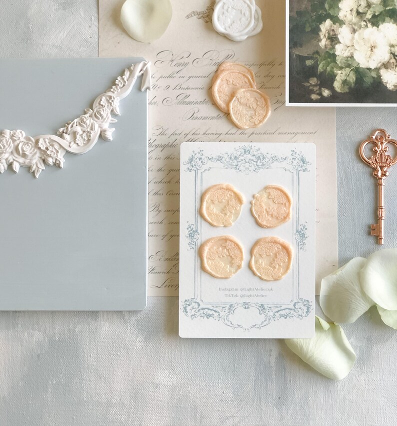 K&ouml;nnte beinhalten: Vier pfirsichfarbene Wachssiegel mit einem floralen Design sind auf einer wei&szlig;en Karte mit einem blauen und wei&szlig;en Rand angeordnet. Die Karte tr&auml;gt den Text "Instagram @light.Melrose TikTok @light.Melrose" aufgedruckt.