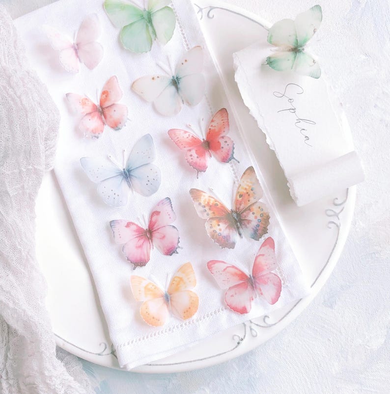 May include: A white fabric napkin decorated with multiple translucent, colourful butterfly cutouts in shades of pink, orange, blue, and green. A matching white paper place card with the name Sophia written in cursive script rests nearby.